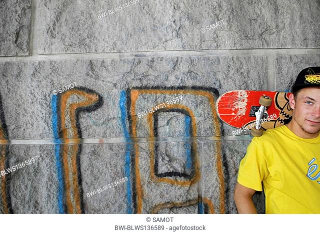male teenager standing in front of a wall with his skateboard on shoulders