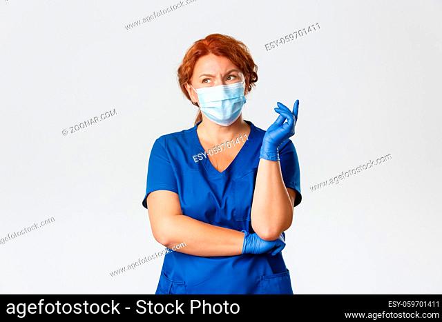 Medical workers, covid-19 pandemic, coronavirus concept. Confused and thoughtful female redhead doctor, nurse in scrubs, face mask and gloves thinking