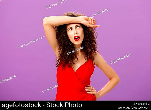 Troubled and tired good-looking european woman. with curly hairstyle in red evening dress wiping sweat of forehead looking at upper right corner perplexed and...