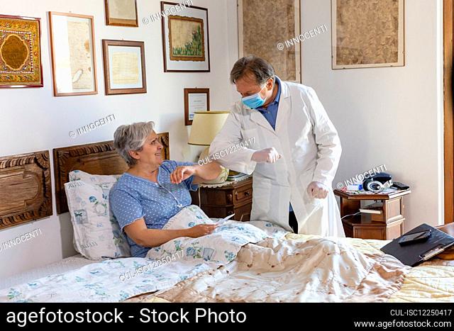 A doctor in a white coat and protective face mask making a home visit to a senior woman patient, greeting by touching elbows
