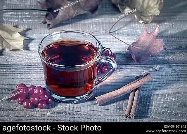 Still life with transparent and fragrant Cup of tea with viburnum and cinnamon on wooden table. Dry autumn maple leaves in the background. Toned photo