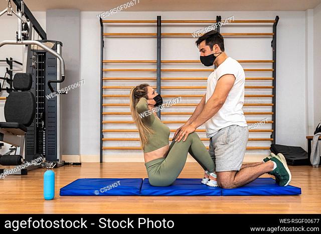 Male athlete with face mask helping female friend while exercising in health club