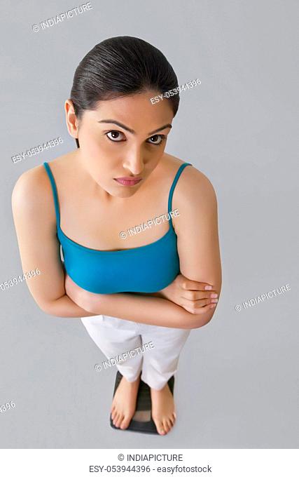 Portrait of worried young woman standing with arms crossed on weighing scale against gray background