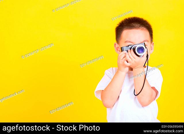 Asian Thai happy portrait cute little cheerful child boy hold photo camera compact doing taking the picture, studio shot isolated on yellow background with copy...