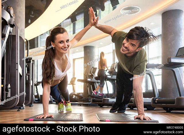 Young fit man and woman smiling and looking at camera while giving high five from basic plank pose during workout at the gym
