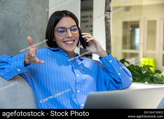 Smiling young female freelancer with the laptop seated on the sofa talking on the cellphone