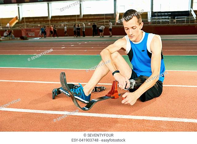 Full length portrait of young amputee athlete sitting on floor in modern gym fixing prosthetic foot, copy space