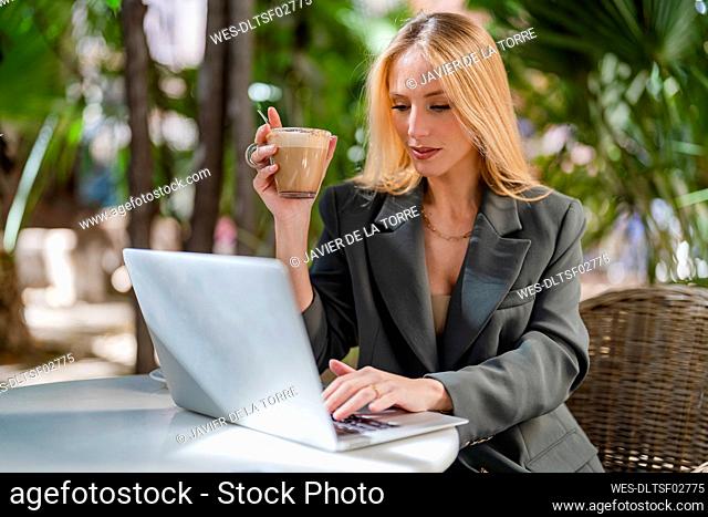 Blond freelancer holding coffee cup using laptop sitting at sidewalk cafe