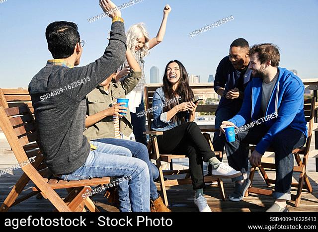 Group of young co-workers hanging out on rooftop patio having a drink and celebrating achievement reached on cell phone