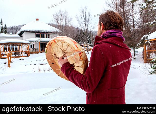 Young man dressed in urban red clothing, pictured from the back is playing his sacred drum with a drumstick. Pictured from the back, outside in winter