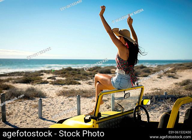 Caucasian woman sitting on beach buggy by the sea wearing straw hat looking toward sea with hands up