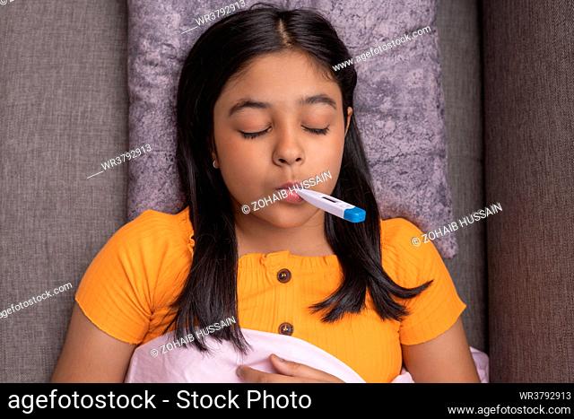 Close-up portrait of a sick girl lying on sofa with a digital thermometer on her mouth