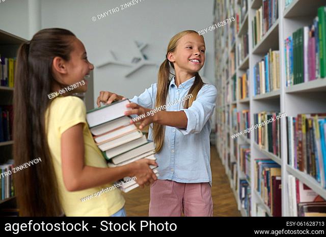 Smiling cute teenage girl helping her cheerful classmate to select textbooks in the reading room