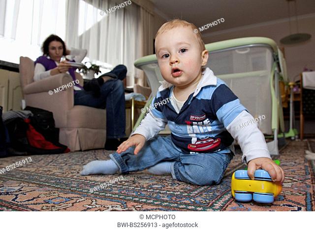 little boy playing on the floor, his mother sitting on a sofa keeping an eye on him