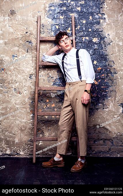 full length fashionable man in white shirt and beige pants with suspenders posing near old wooden ladder on brick wall background, standing and looking away