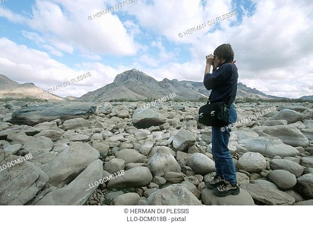 Woman looking through binoculars on rocky river bed, Pella, Northern Cape Province, South Africa