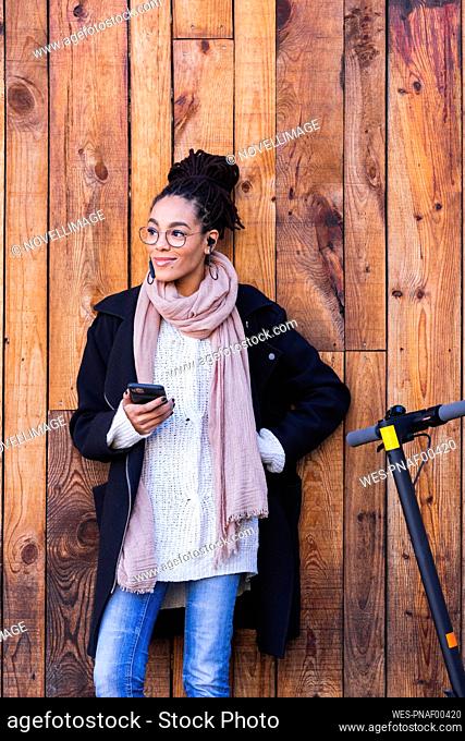 Young female millennial holding smart phone looking away while leaning against wooden wall by electric push scooter
