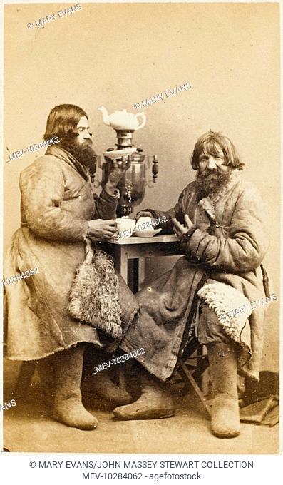 Two Russian peasants in thick sheepskin skin overcoats sit at a table drinking tea from a samovar. Both men are wearing 'valenki' - thick felt winter boots
