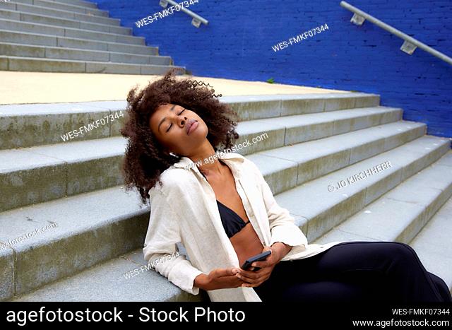 Young Afro woman with eyes closed listening music through in-ear headphones sitting on steps