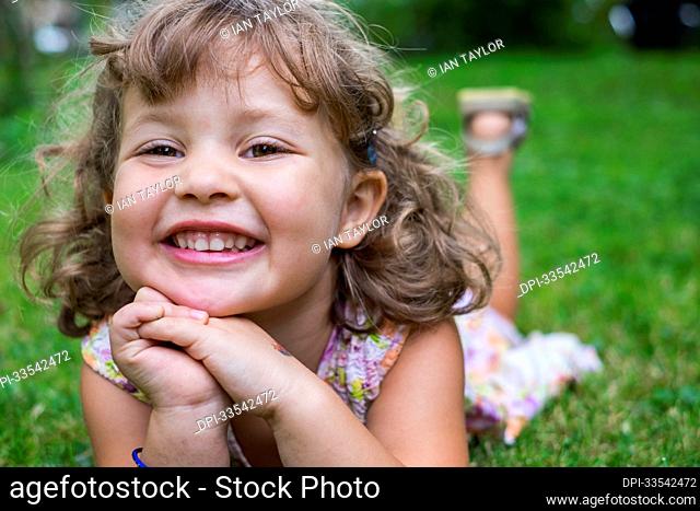 Portrait of a preschooler girl with blond curly hair and brown eyes, lying on the grass with hands folded in front of her; Toronto, Ontario, Canada