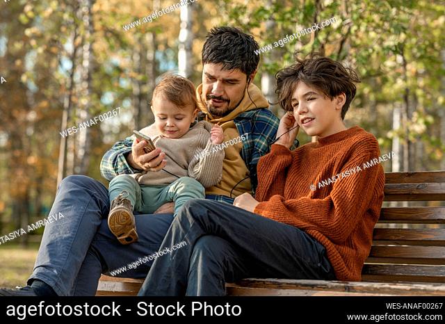 Man with son on lap holding mobile phone listening to music through in-ear headphones sitting by boy on bench