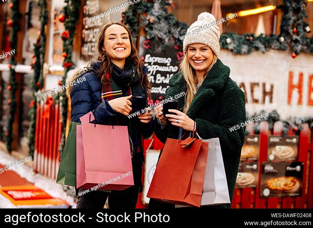 Amigos alegres con bolsas de compras disfrutando del vino caliente en el mercado navideño