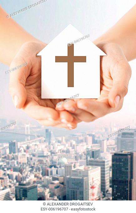 religion, christianity and charity concept - close up of woman hands holding paper house with christian cross symbol over city background