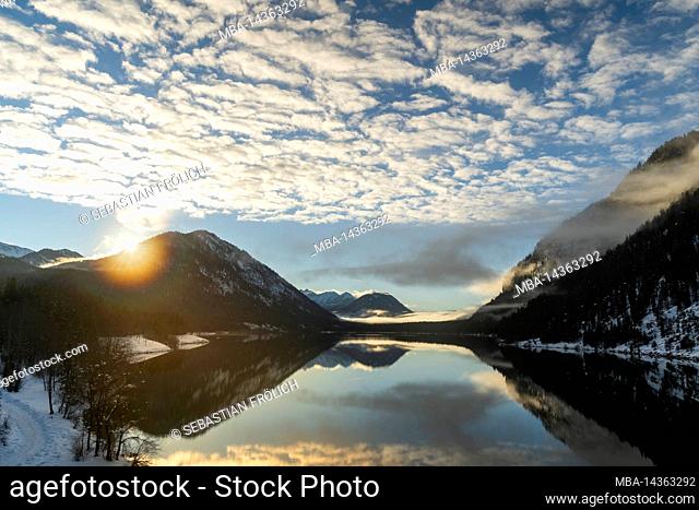 The evening sun in winter on the shore of the Sylvenstein reservoir. Inetressant clouds and fog atmosphere as a reflection in the clear water of the alpine lake...