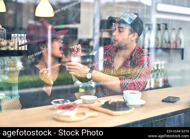 Pretty interracial couple sits in a cafe. They are eating desserts. White guy in a red shirt feeds his black girlfriend in a crimson hat. Horizontal