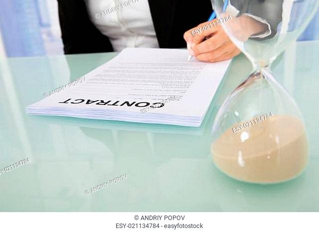 Businesswoman Signing Contract With Hourglass At Desk