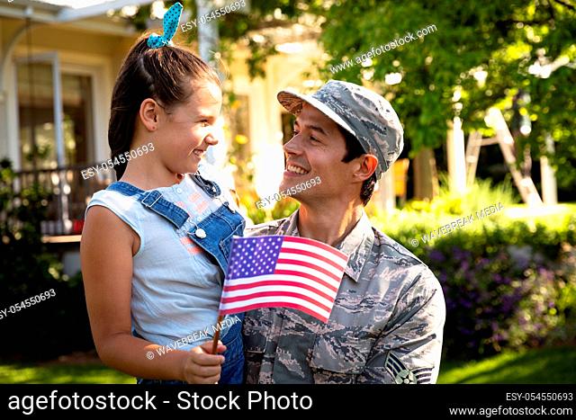 Front view of a young adult mixed race male soldier in the garden outside his home, carrying his daughter, who is holding a US flag, both smiling at each other