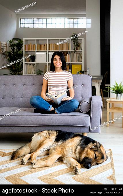 Smiling woman with book sitting on sofa while dog sleeping on carpet in living room