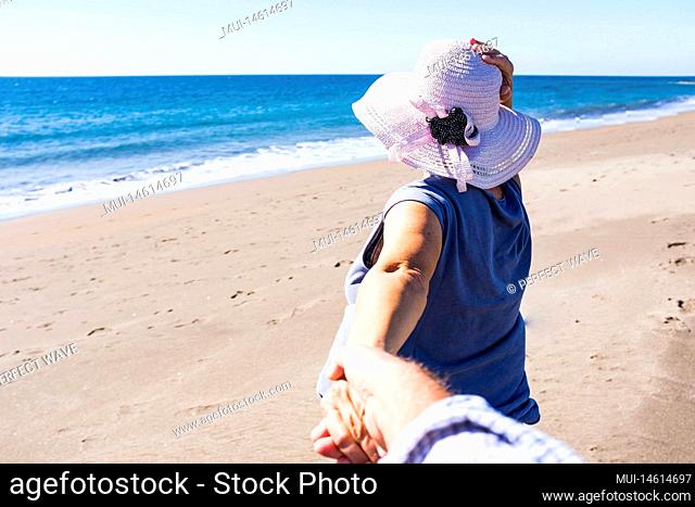 one senior and mature woman walking and holding the hand of her husband at the beach on the sand - summertime and discovering ew places together - pensioner...