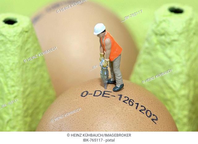 Miniature man wearing a hardhat drilling into a stamped egg in a carton of eggs