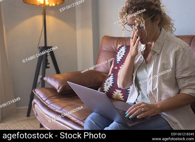 Beautiful young woman rubbing eye while working on laptop sitting on sofa and smiling. Tired businesswoman using laptop in the living room at home