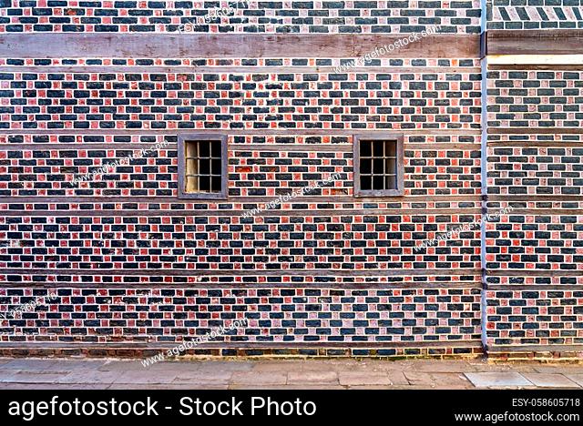 Wall with black and red bricks with white seam and two small adjacent windows with metal bars on cobblestone road
