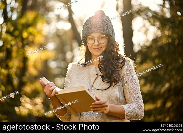 beautiful woman in a cap and glasses is holding a book in an autumn forest in hands