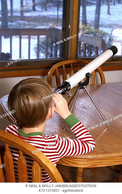 Little boy inside looking outside in winter through a telescope
