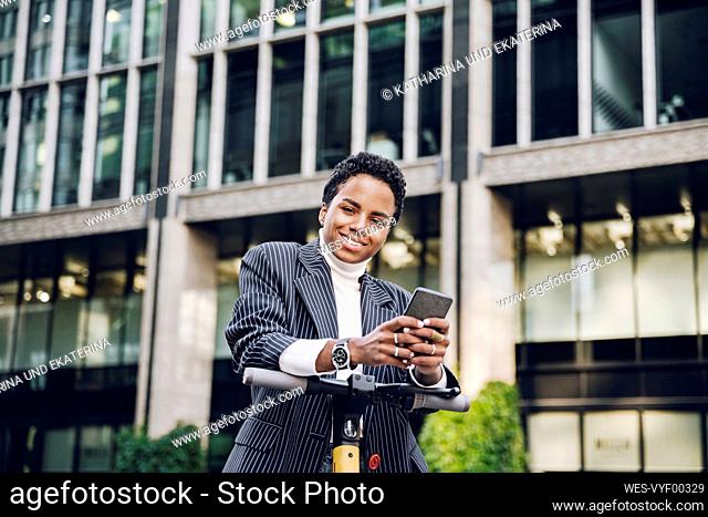 Smiling businesswoman holding mobile phone leaning on handlebar of electric push scooter in city