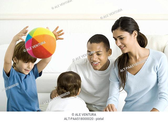 Family gathered around toddler girl, smiling, boy throwing ball in the air