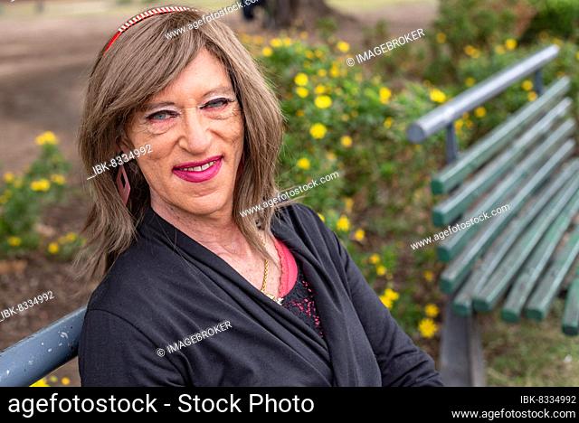 Transgender man with make-up on and wearing womans clothes sitting on a bench in a public park