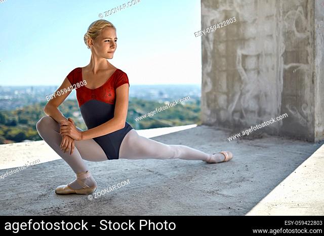 Beautiful ballerina is posing on the concrete floor of the unfinished building on the cityscape background. She wears a red-gray leotard with light leggings and...