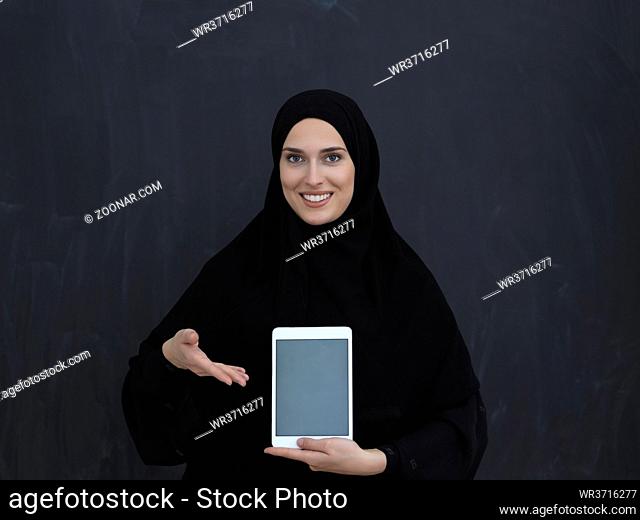 Young Arab businesswoman in traditional clothes or abaya and glasses showing tablet computer display in front of black chalkboard