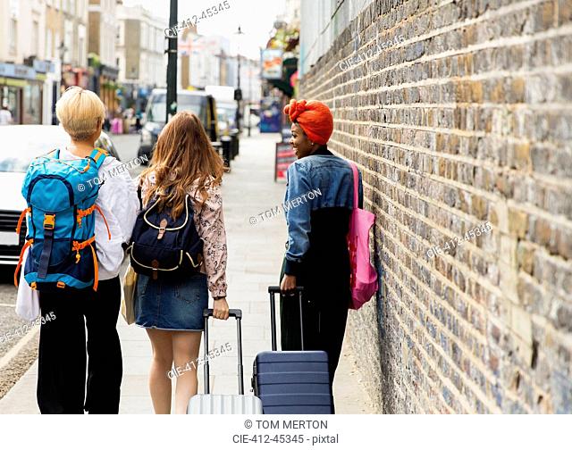 Young women friends with suitcases and backpack walking on urban sidewalk