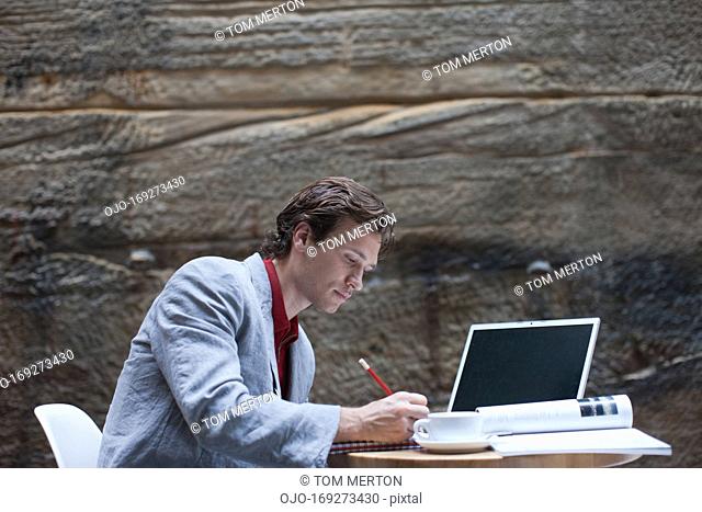 Businessman writing in notepad at table with laptop and coffee