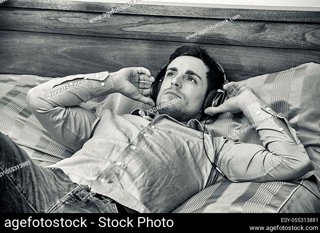 Handsome young man in bed listening to music with headphones, using cell phone or MP3 player