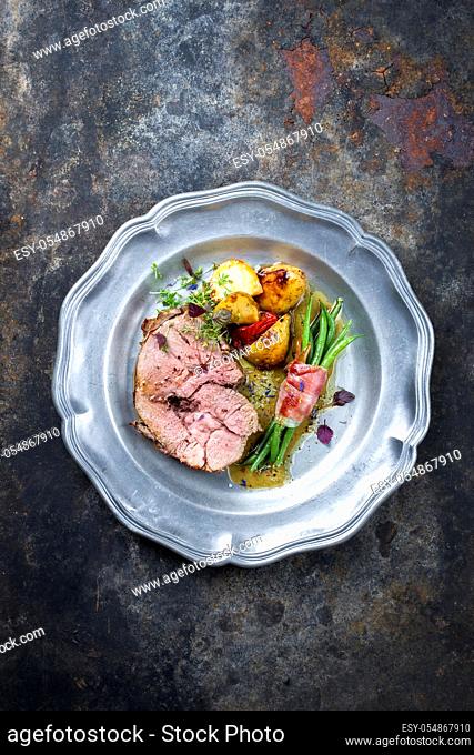 Barbecue marinated Greek lamb roast with potatoes and beans rolled in bacon as top view on a pewter plate