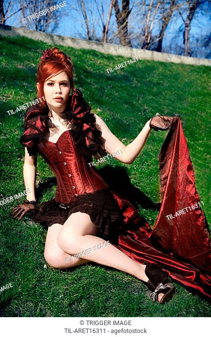 Redhead in Victorian dress sitting on grass, holding the hem of the dress