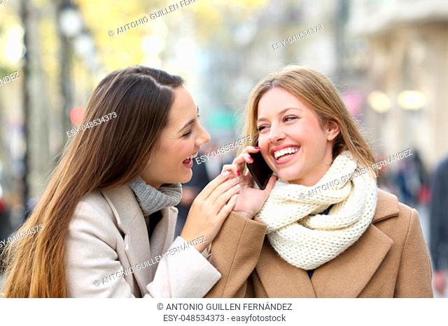 Two happy friends talking on mobile phone on the street in winter