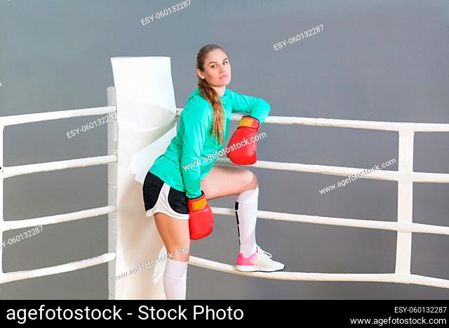 Beautiful athletic young woman with collected hair wearing in green long sleeve and red gloves standing, leaning on near boxing ring and looking at camera